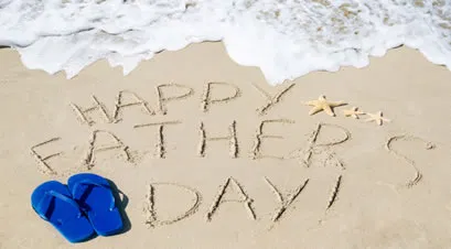 Happy Father’s Day written in beach sand beside blue thongs