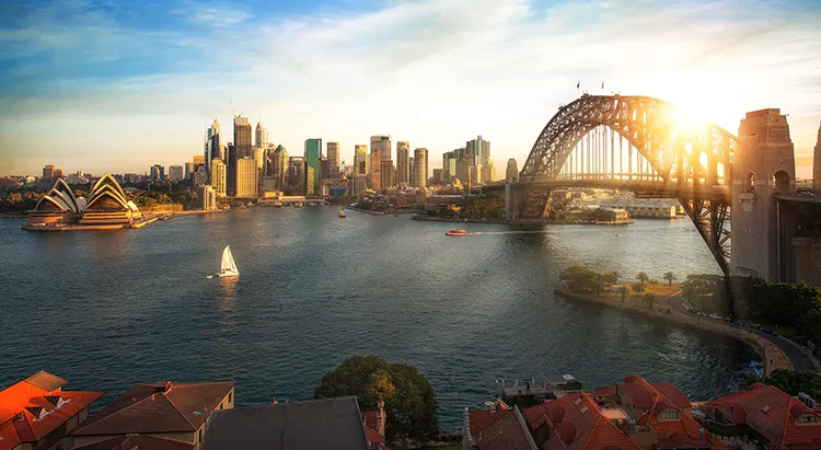 Sydney Harbour at sunset seen over the rooftops of waterfront homes, with the Opera House, city skyline and Harbour Bridge beyond